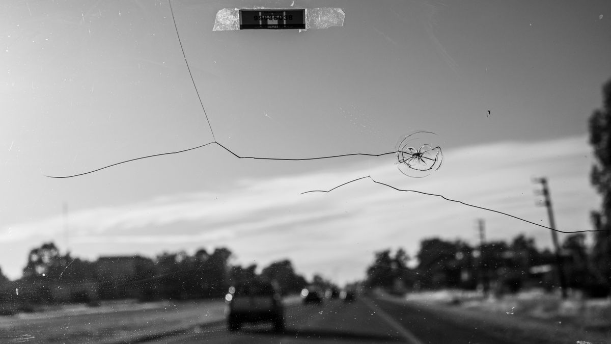 Pare-brise de voiture marqué d'un impact étoilé prolongé par une longue fissure, vu depuis l'habitacle sur une route de campagne