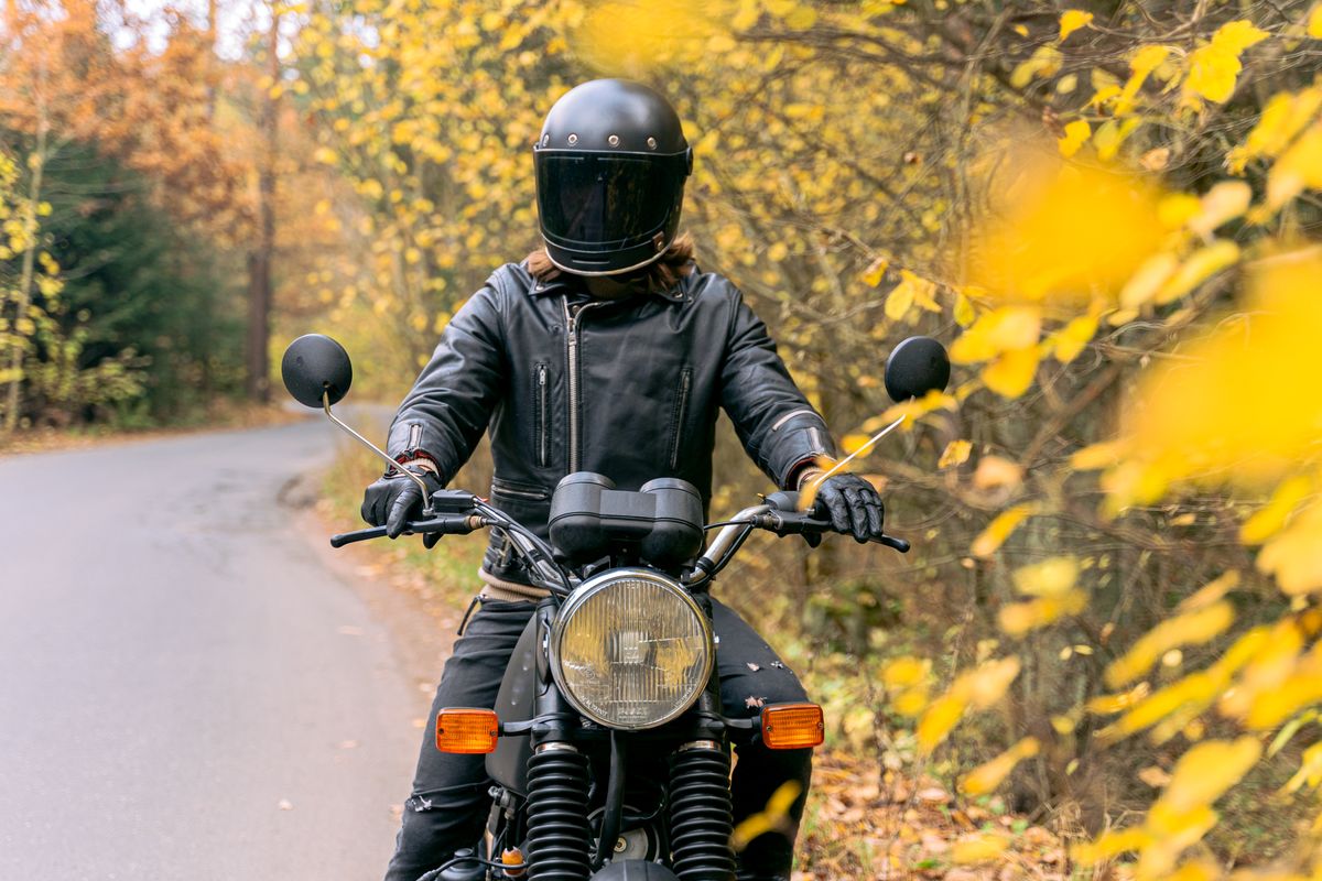Motard en blouson de cuir, casque intégral et gants, à l'arrêt sur une route de campagne en automne