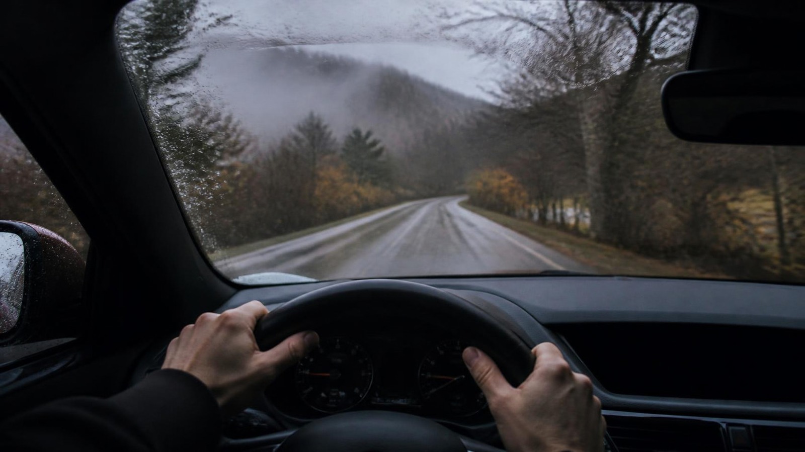 Mains d'un conducteur sur le volant d'une berline, route de campagne mouillée vue à travers le pare-brise en automne