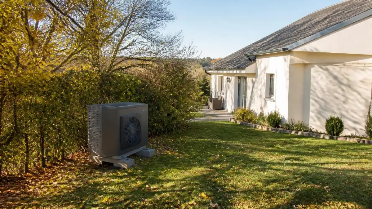 Unité extérieure d'une pompe à chaleur air-eau posée sur des plots dans le jardin d'une maison, en fin d'après-midi d'automne